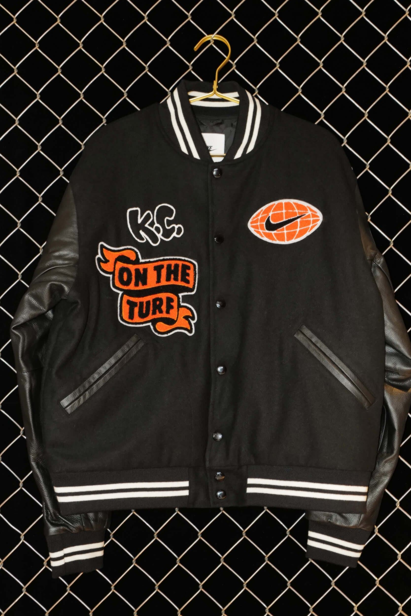A black varsity jacket with “On the Turf” and Nike football patches, hung against a chain-link fence