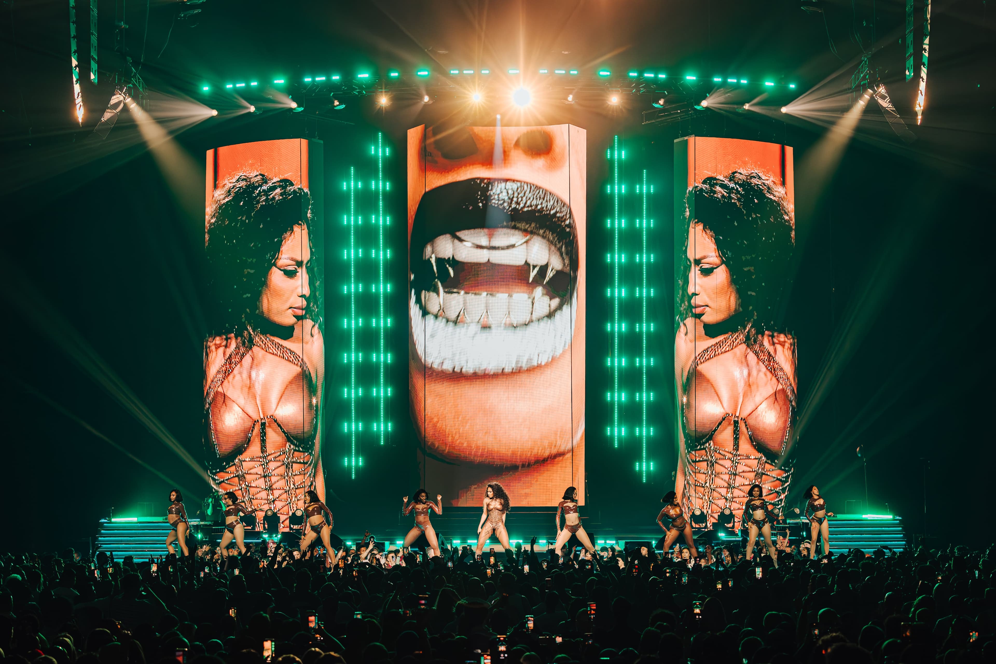 A fanged mouth on the centre screen between portraits of the artist, dancers on the green-lit stage below