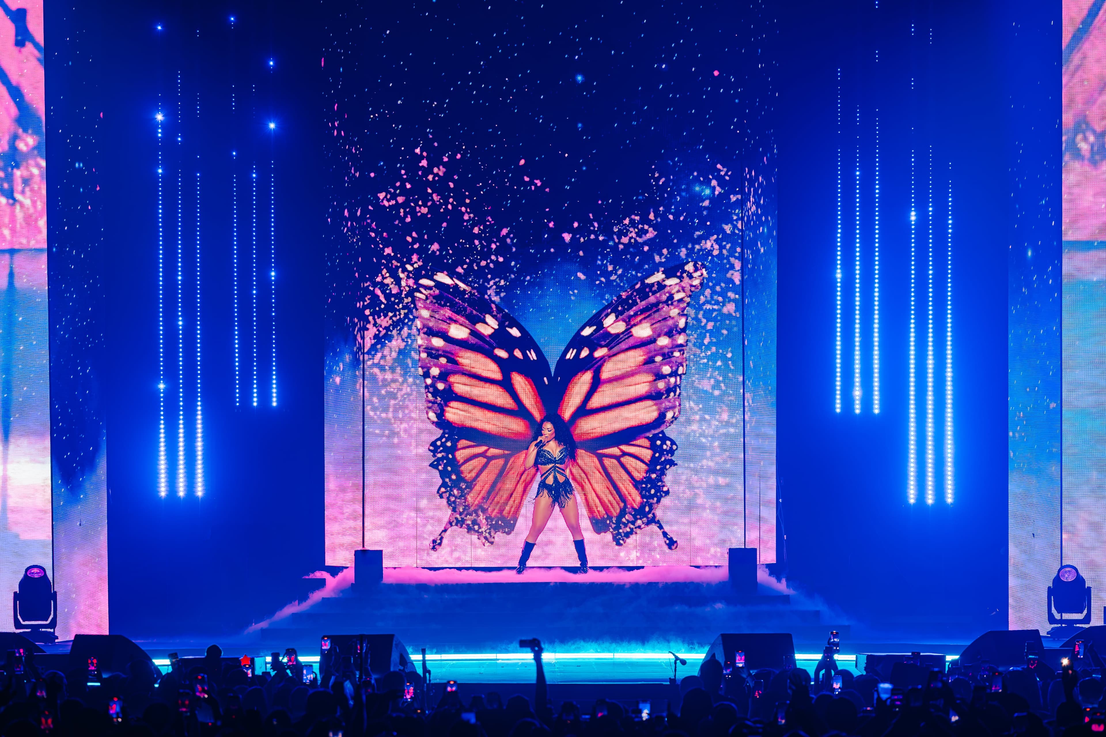 The artist in giant orange butterfly wings on the blue-lit stage, sparks rising on the screens behind