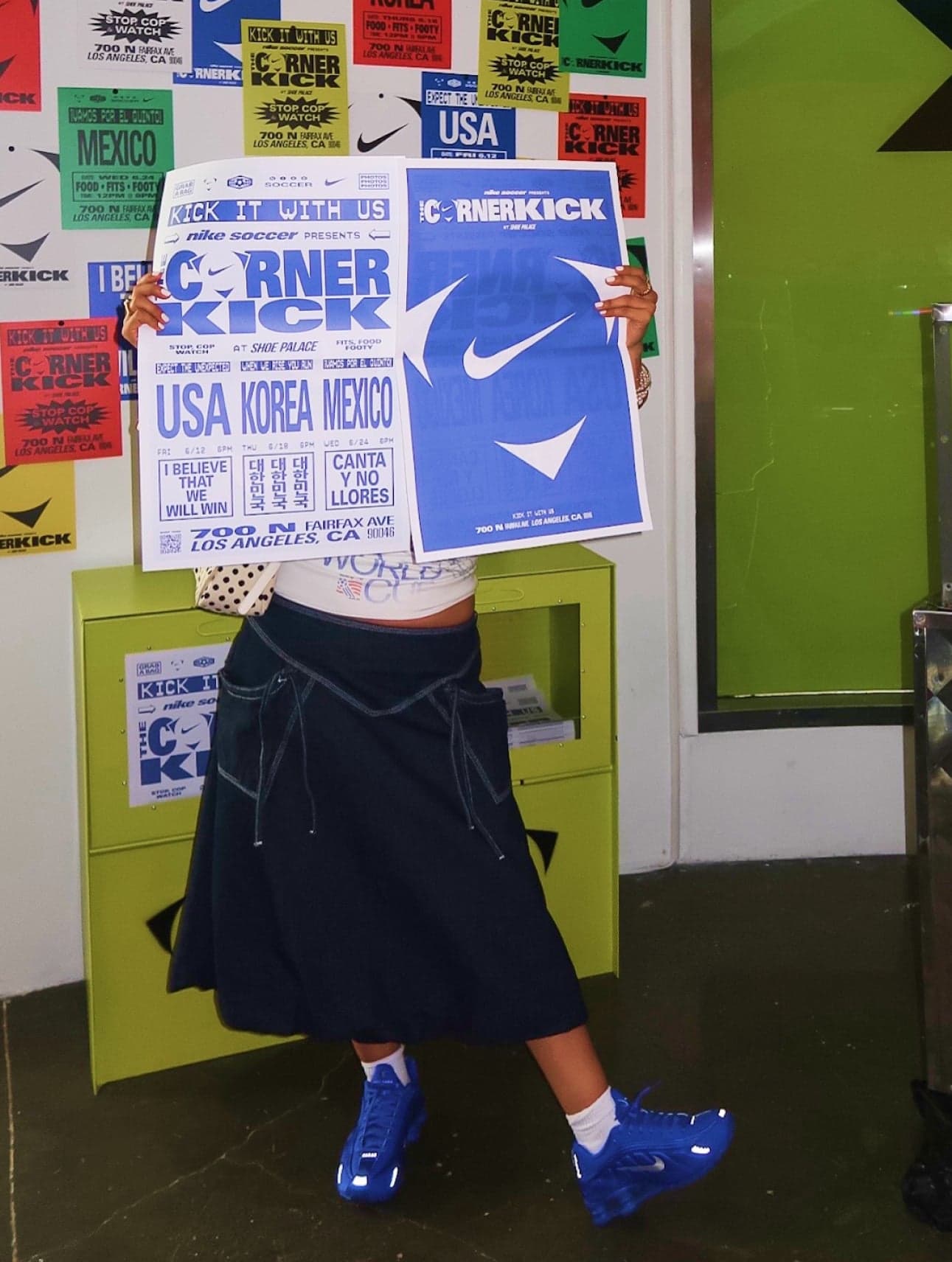 A guest in a black skirt and blue sneakers holding up two Corner Kick match posters in front of a sticker wall