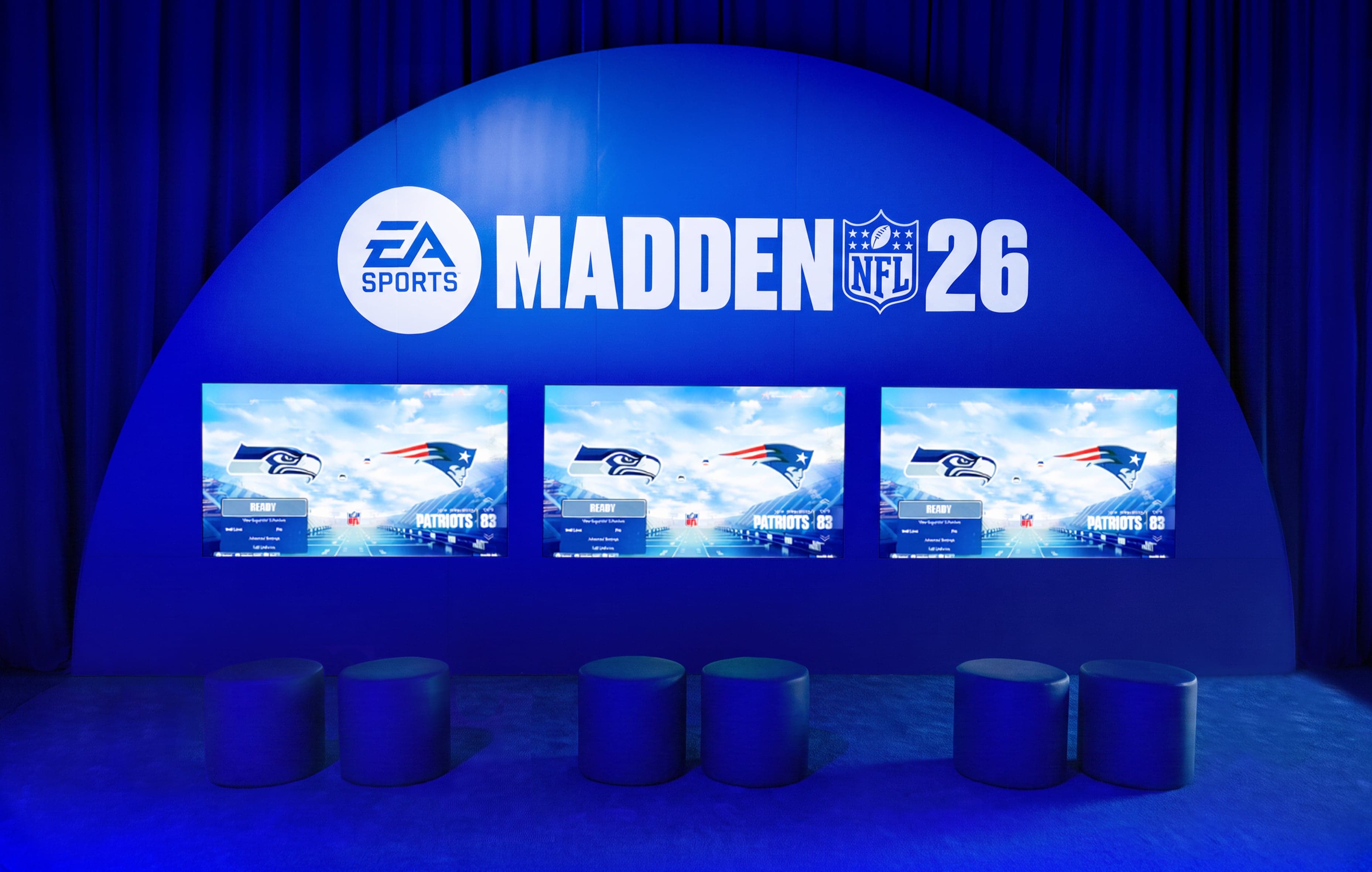The Madden NFL 26 gaming lounge: a blue dome over three screens showing Seahawks vs Patriots, with round stools in front