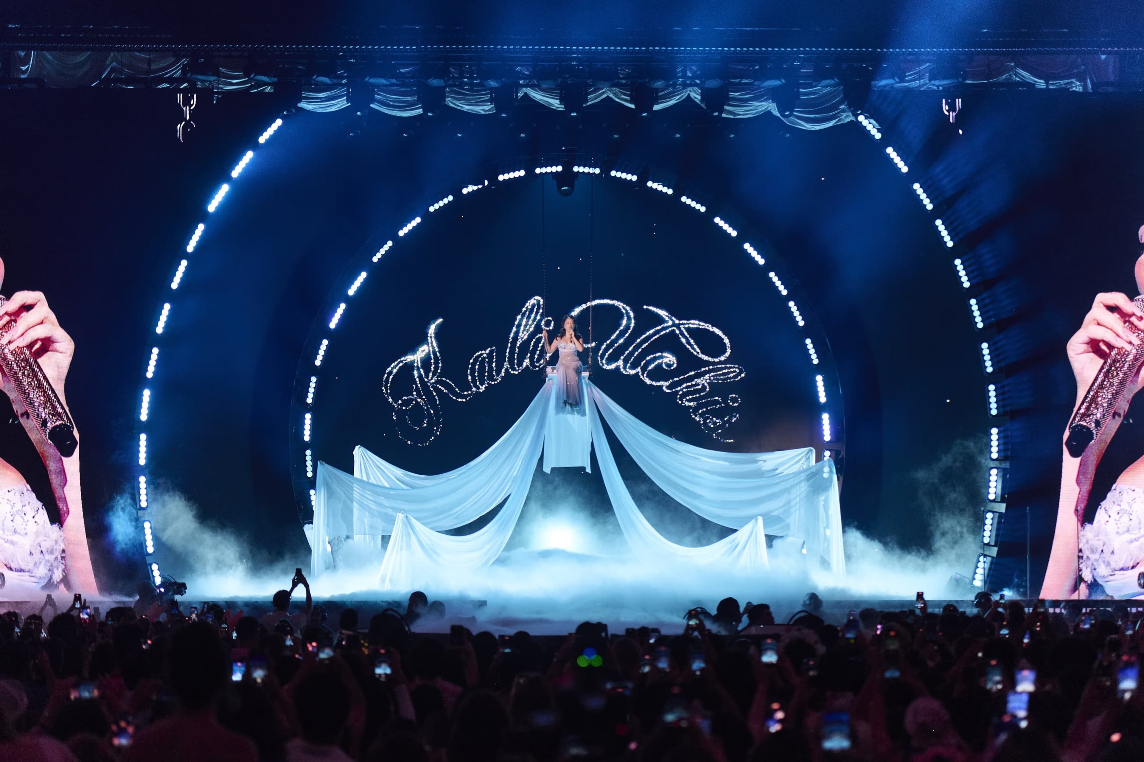 A singer in a long white gown spread across the stage under the artist’s name in script, lit blue
