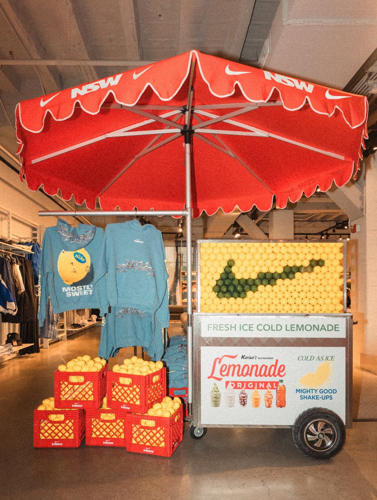 A red scalloped umbrella over a lemonade cart stacked with crates of lemons, with blue lemon-print hoodies hanging behind it