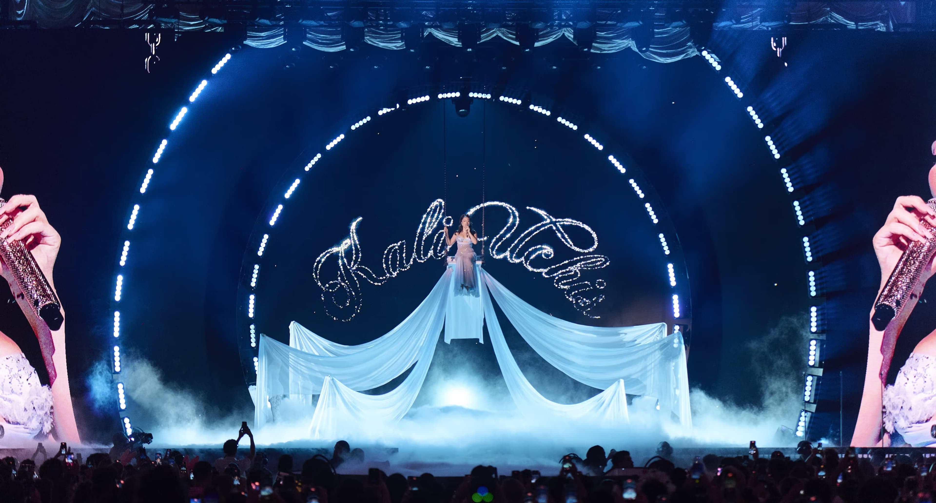 A singer in a long white gown spread across the stage under the artist’s name in script, lit blue