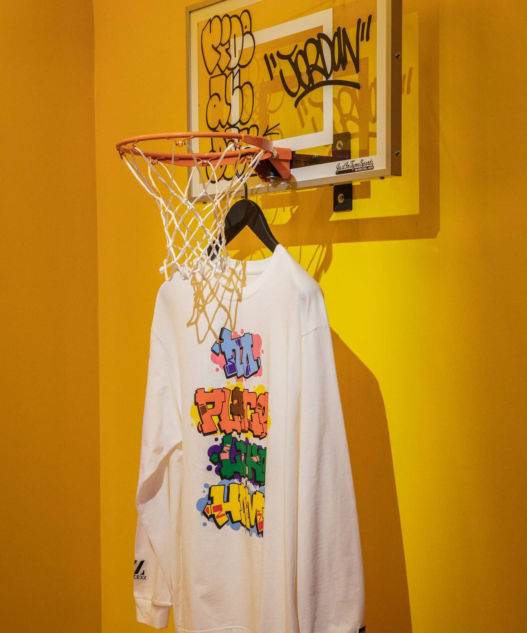 A white graphic hoodie hanging from a basketball hoop on a yellow wall