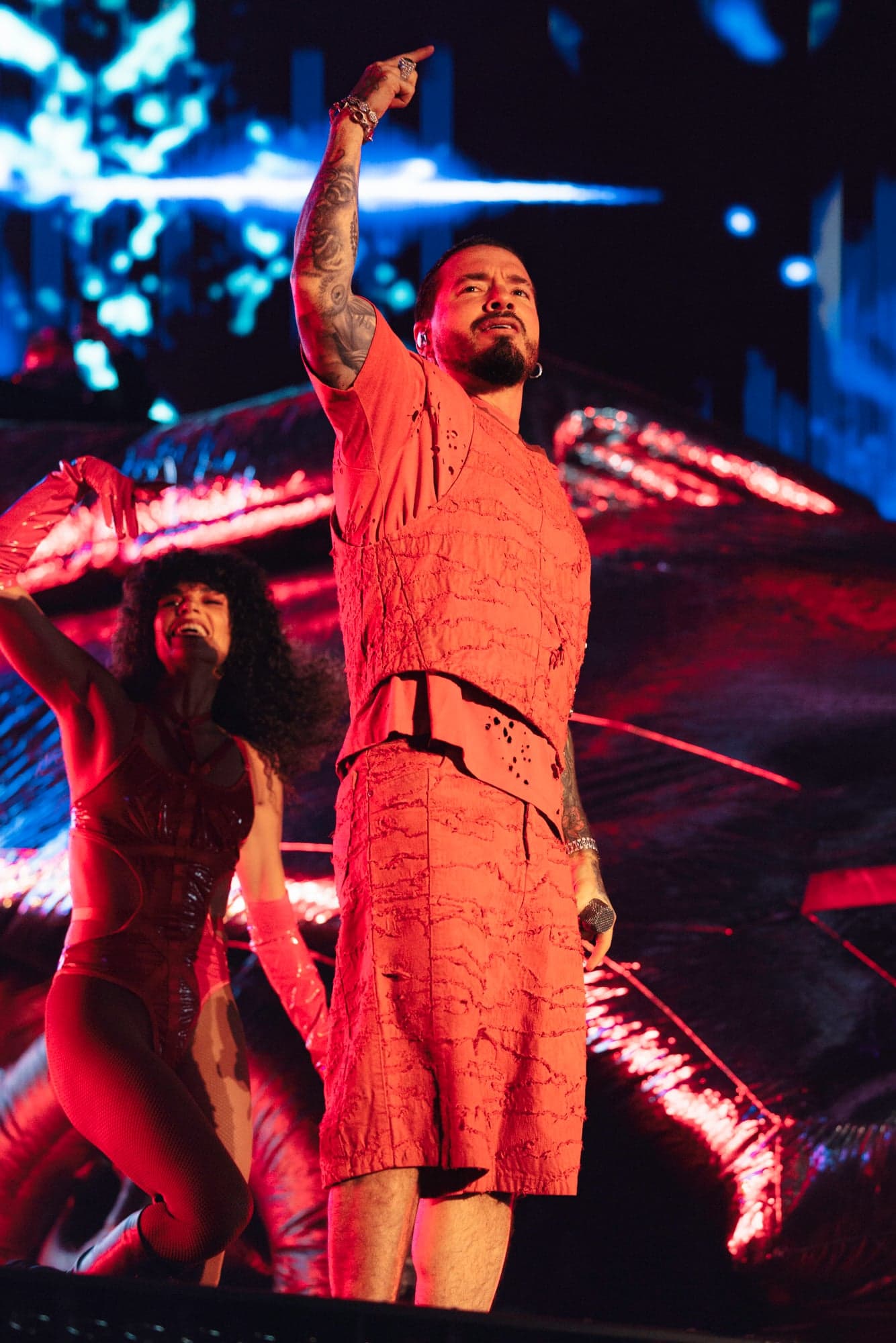 A performer in a terracotta outfit raising an arm on stage, with a dancer behind
