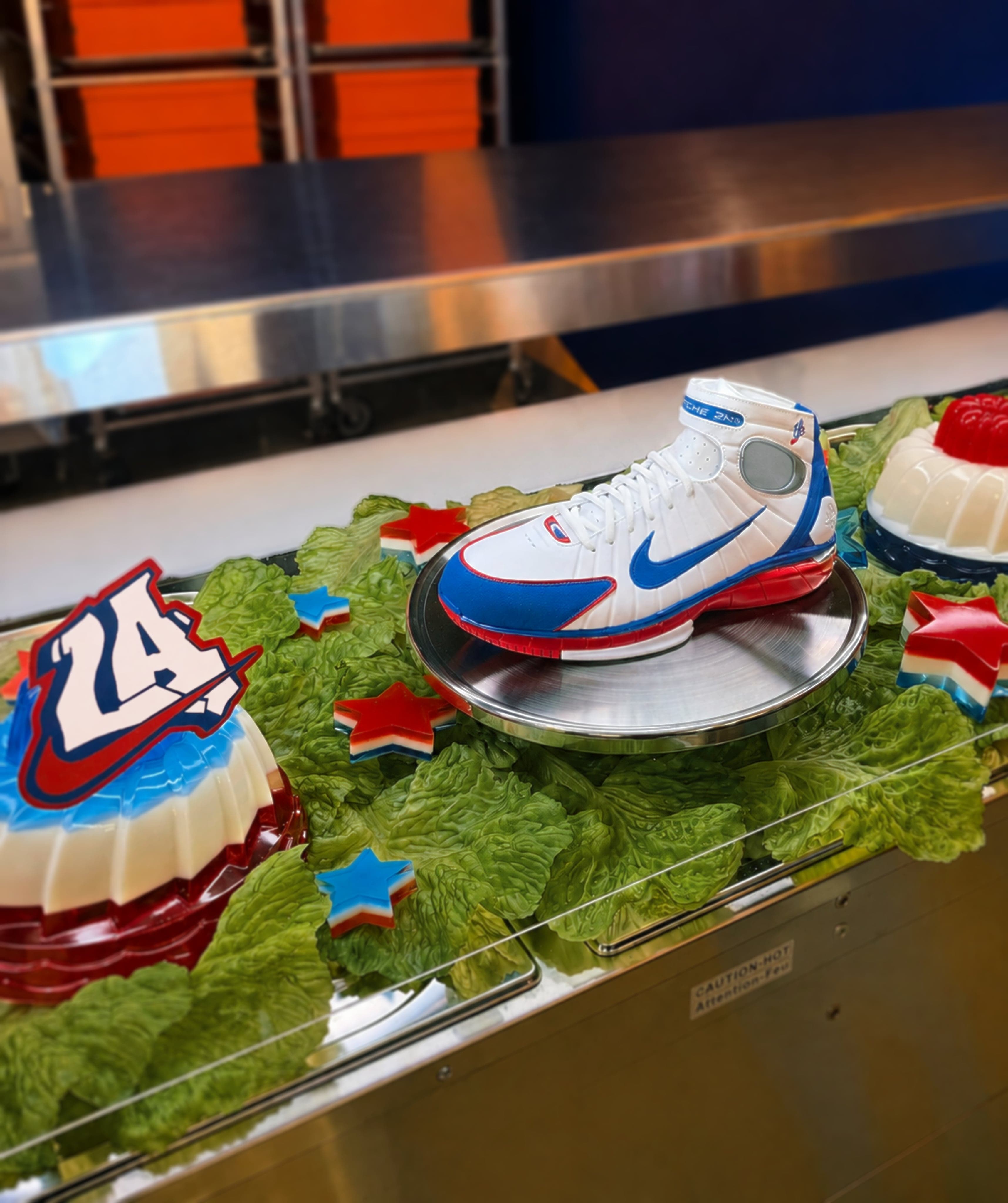 A sneaker served like a cafeteria dish on green jelly and grass, beside a cake topped with the number 24