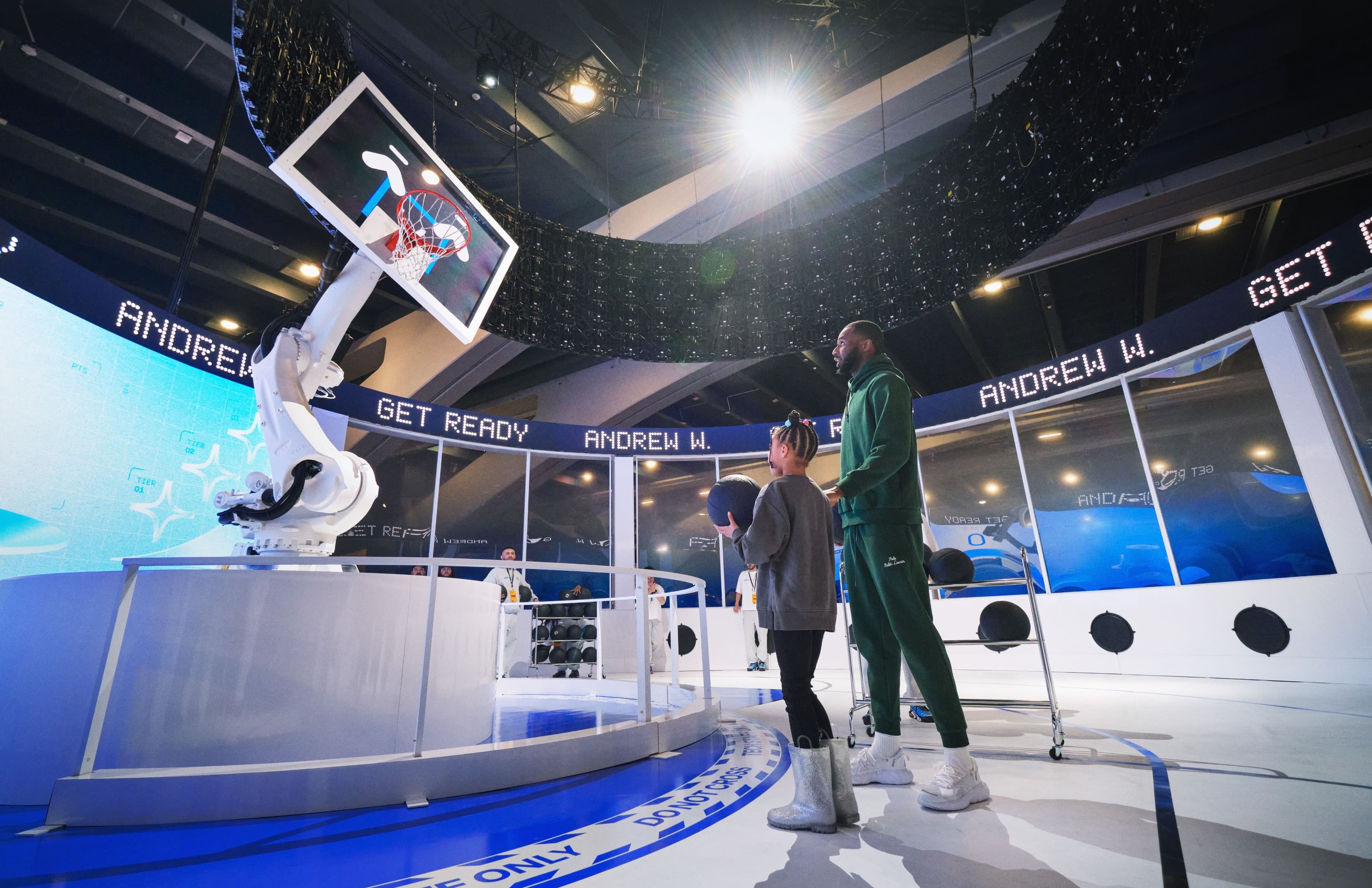 A child and an adult watch the robotic hoop move as their name appears on the “Get Ready” ticker