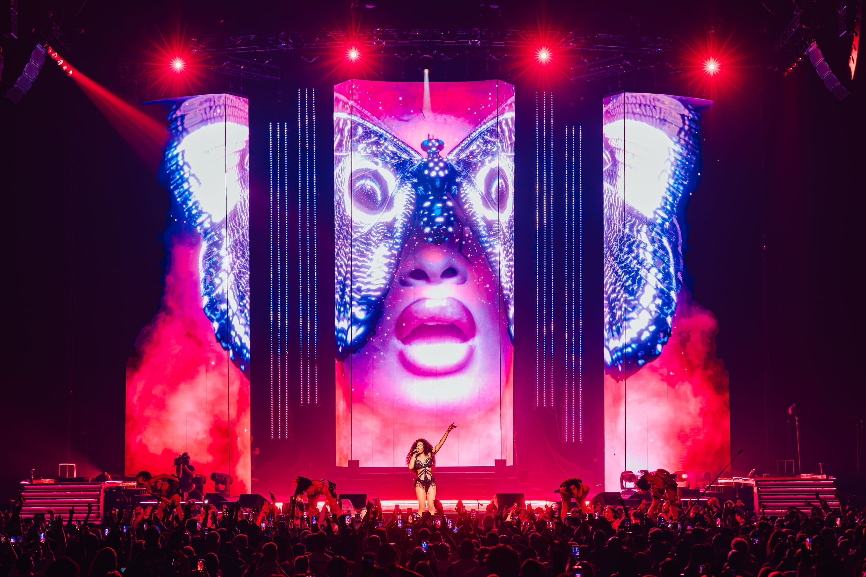 The artist on stage under red lights, between screens showing a face masked by butterfly wings