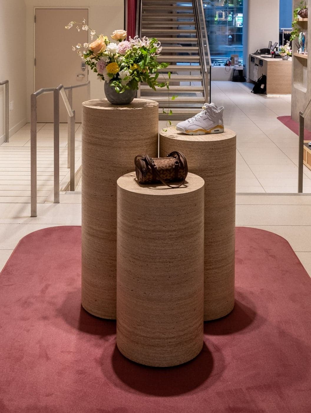 Wooden cylinder pedestals displaying a sneaker, a bag and flowers on a pink rug