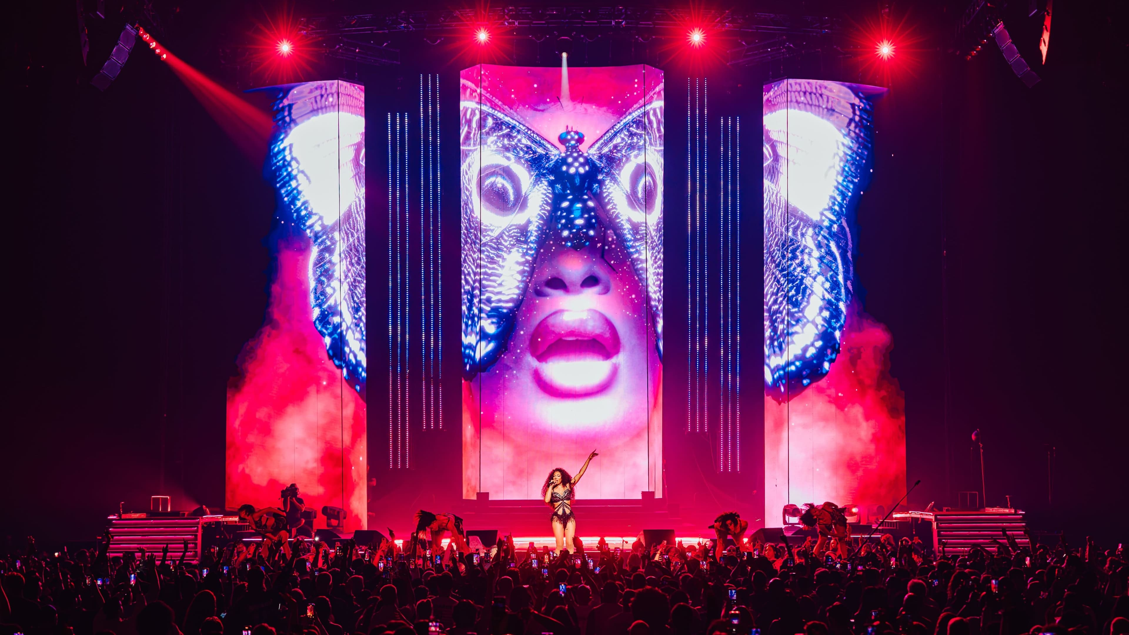 The artist on stage under red lights, between screens showing a face masked by butterfly wings