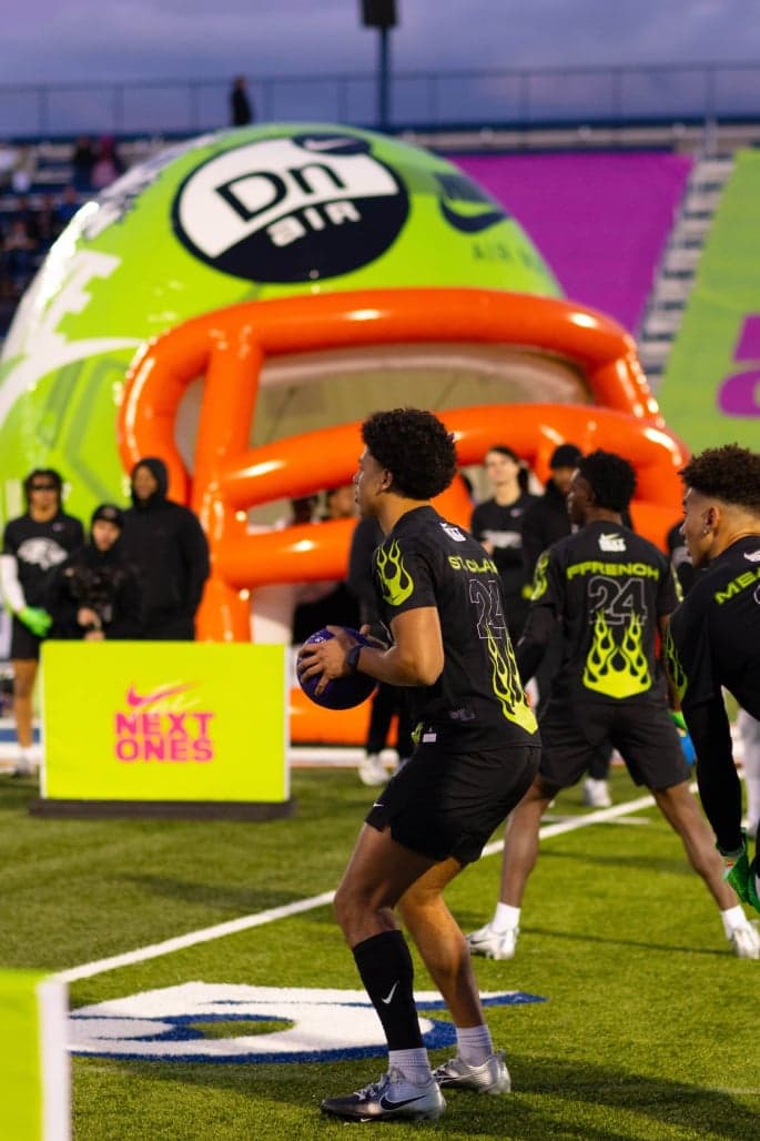 Players running drills in front of the inflatable helmet tunnel and a “Next Ones” banner
