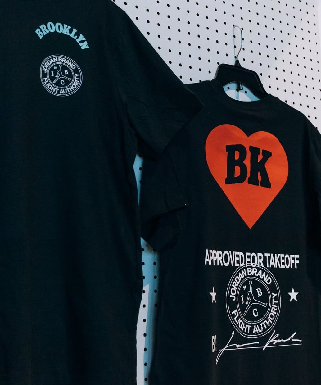 A black T-shirt with a Jordan Brand Classic seal and a black BK heart T-shirt hanging on a pegboard wall