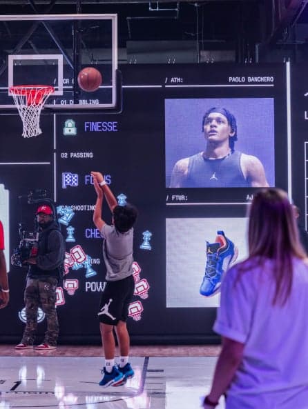A young player shooting on the court in front of a big screen showing his stats and a player portrait