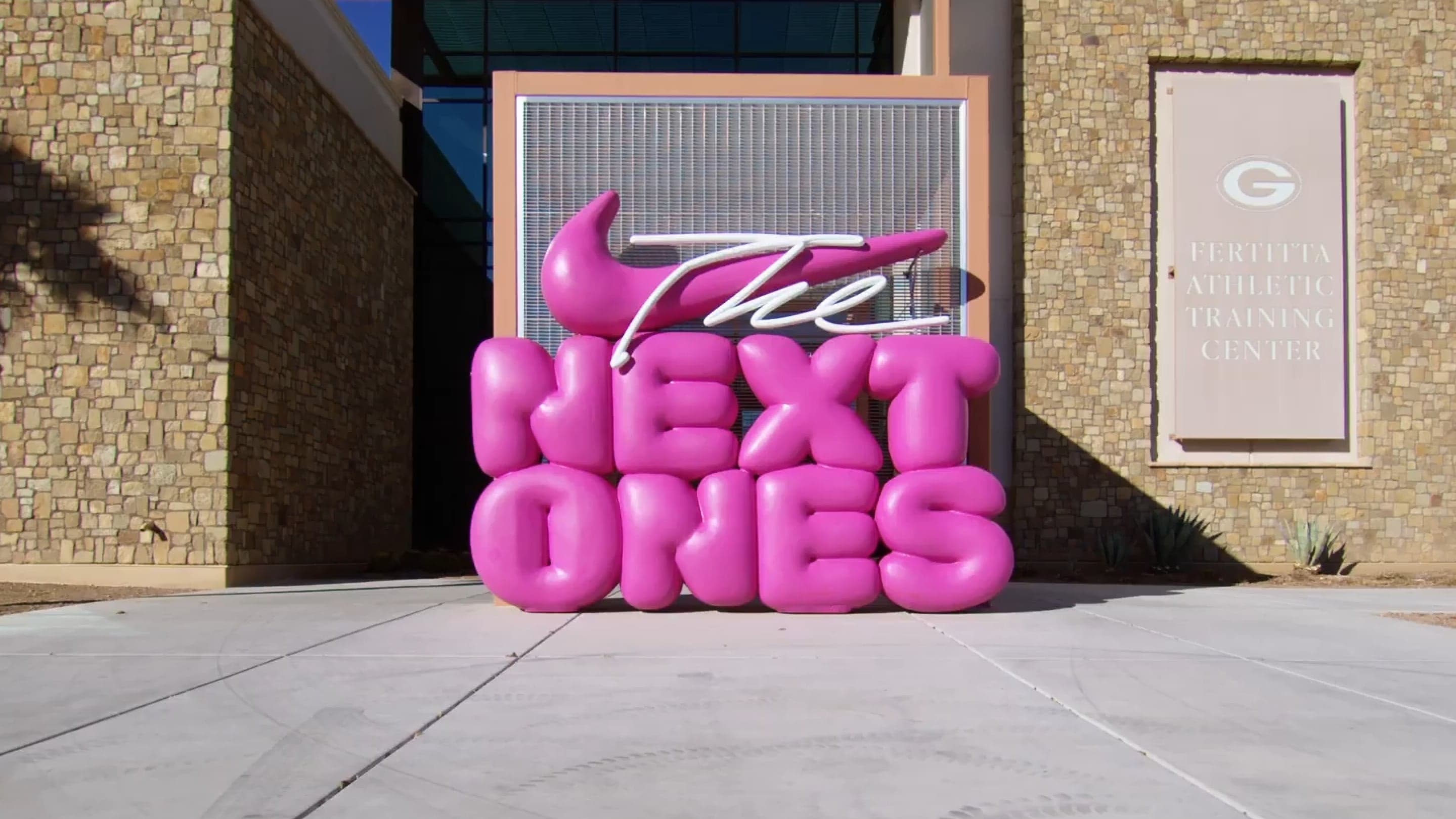 The pink inflatable “The Next Ones” sculpture with a Nike swoosh outside a training center entrance