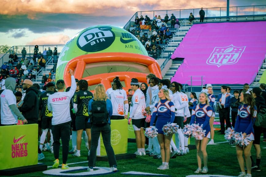 Fans and cheerleaders gathered on a football field around an inflatable helmet, with a purple NFL banner behind them