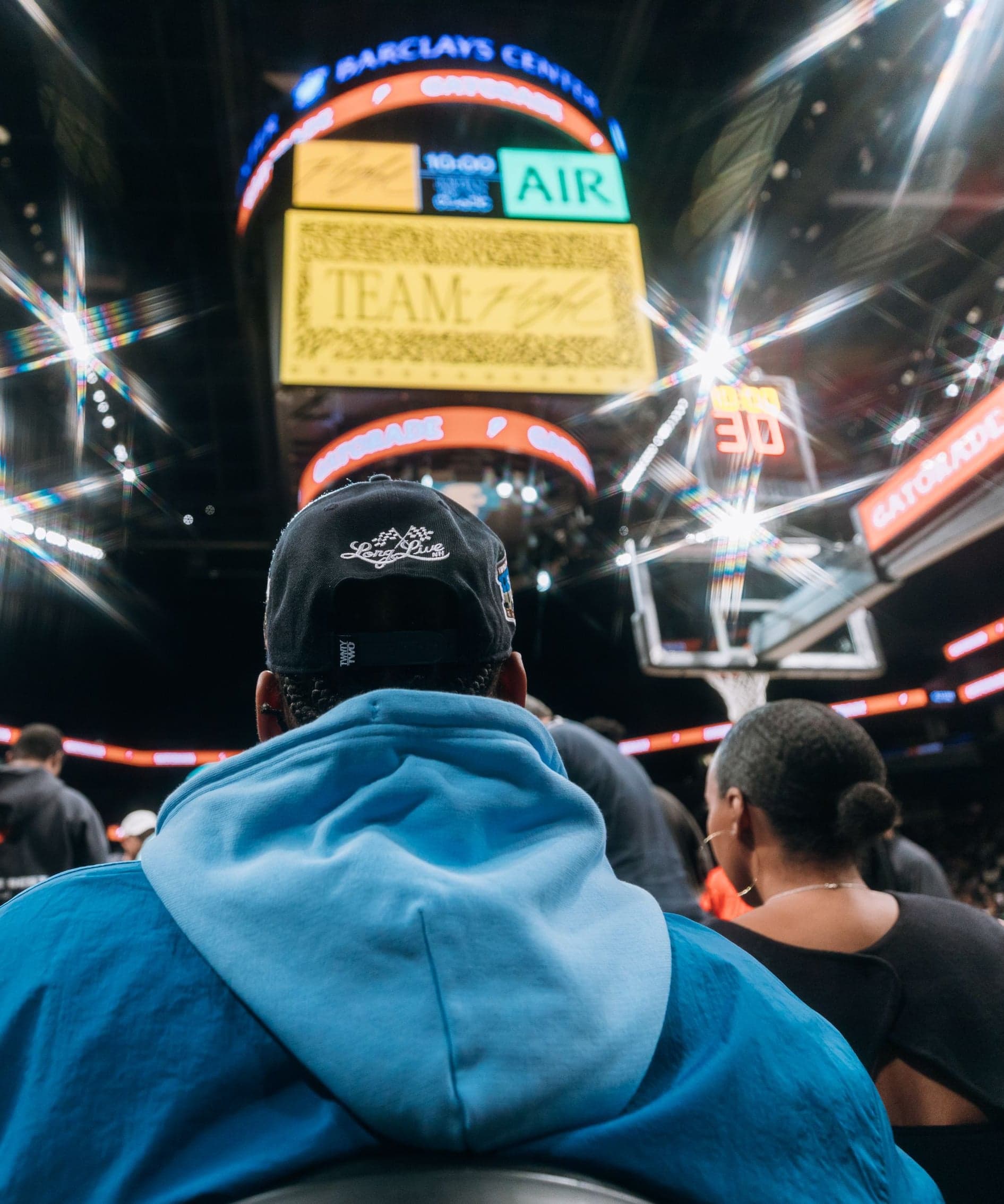 A fan in a blue hoodie and cap watching the game beneath the arena scoreboard