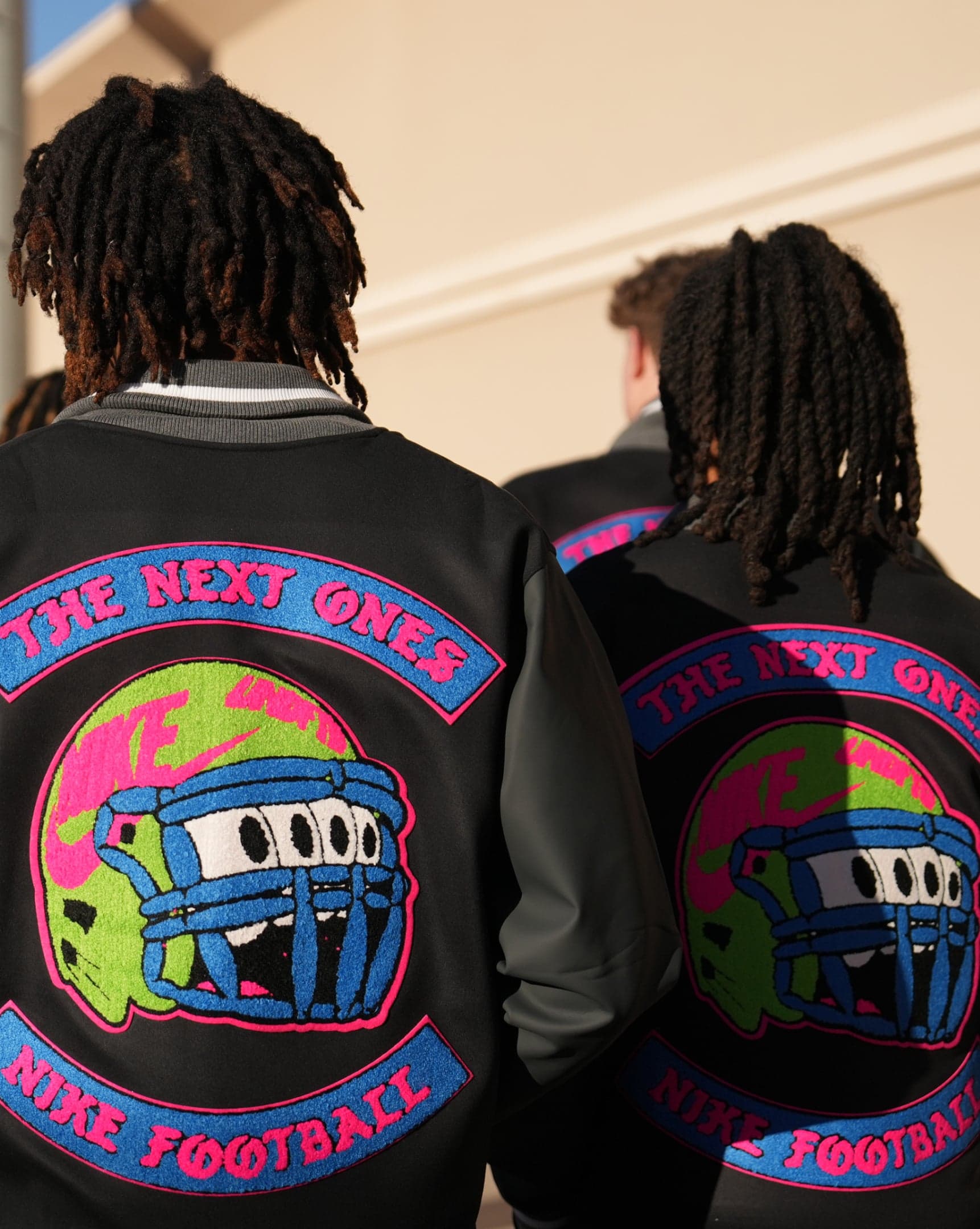 Two players seen from behind in black varsity jackets with “The Next Ones” helmet patches