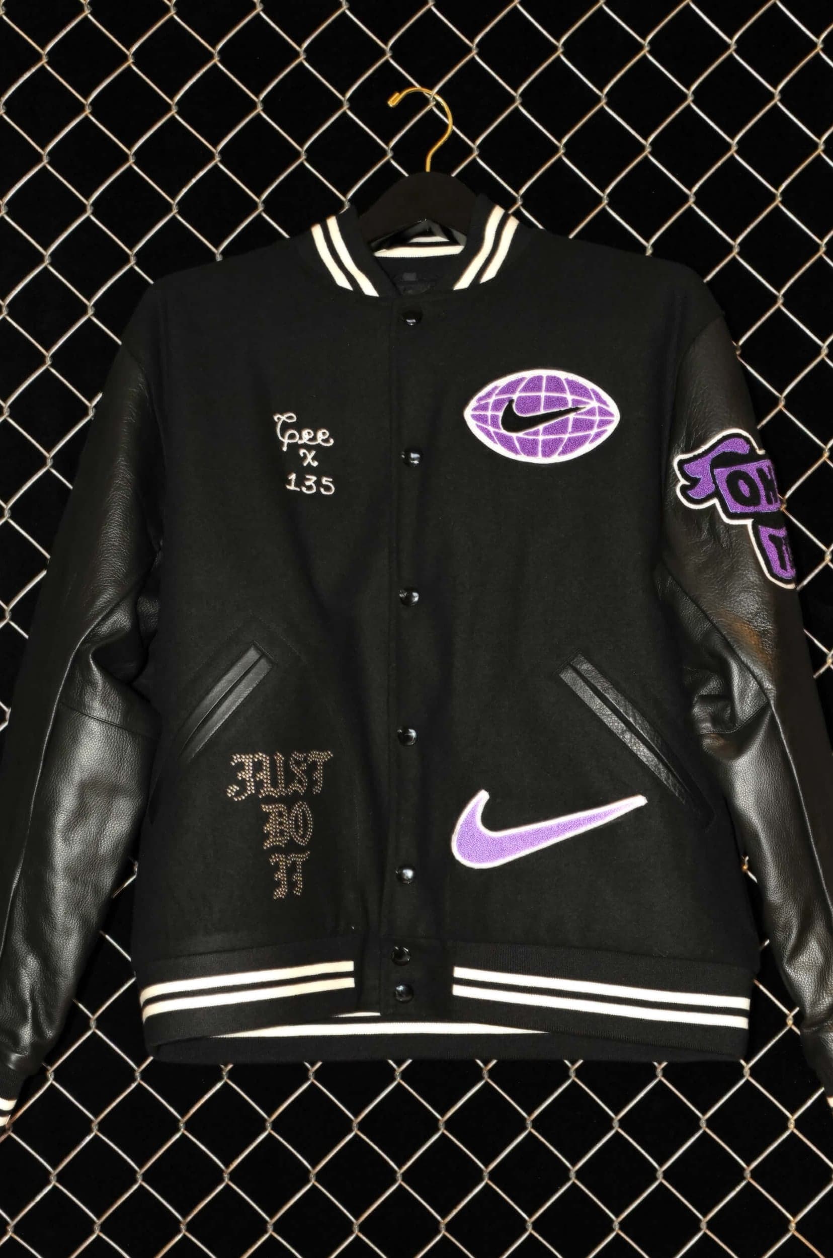 A black varsity jacket with purple Nike patches and chain-stitched lettering, hung against a chain-link fence