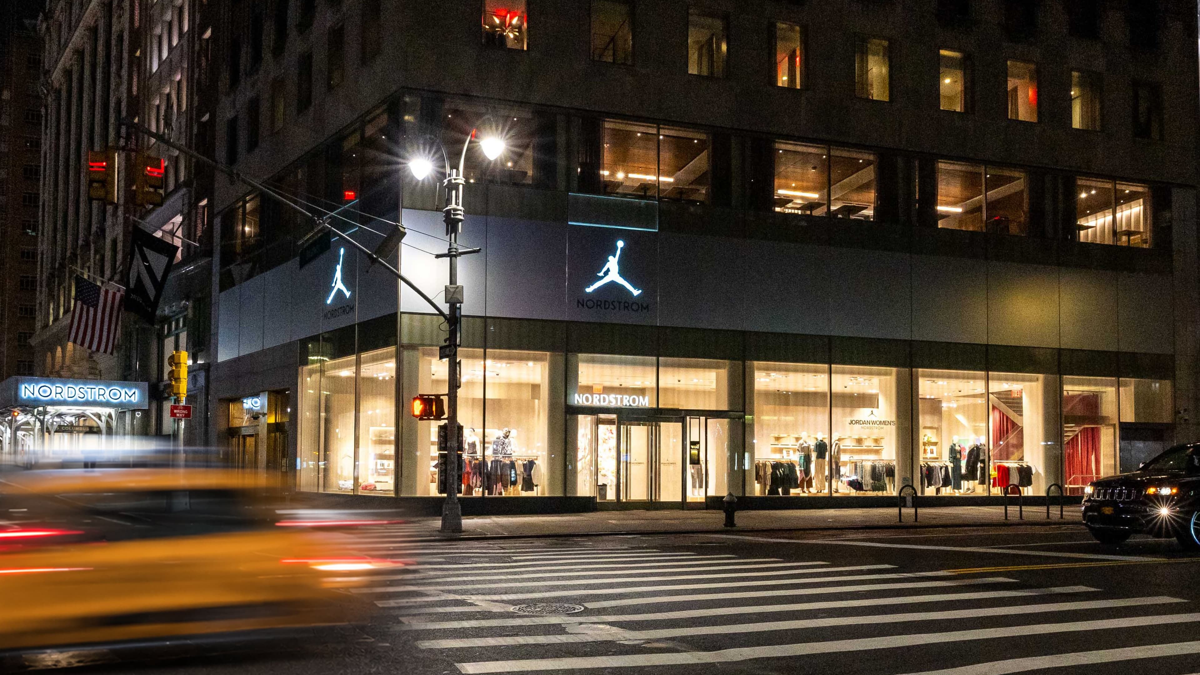 The Nordstrom corner in Manhattan at night, Jumpman logos lit above the windows as a taxi blurs past