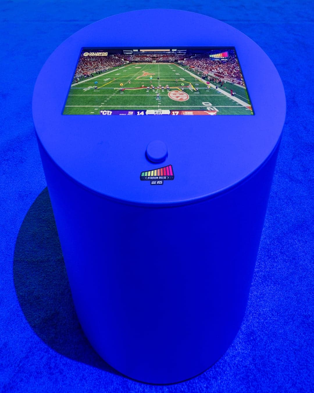 A blue cylindrical podium with a touchscreen showing a college football game and a crowd-noise meter
