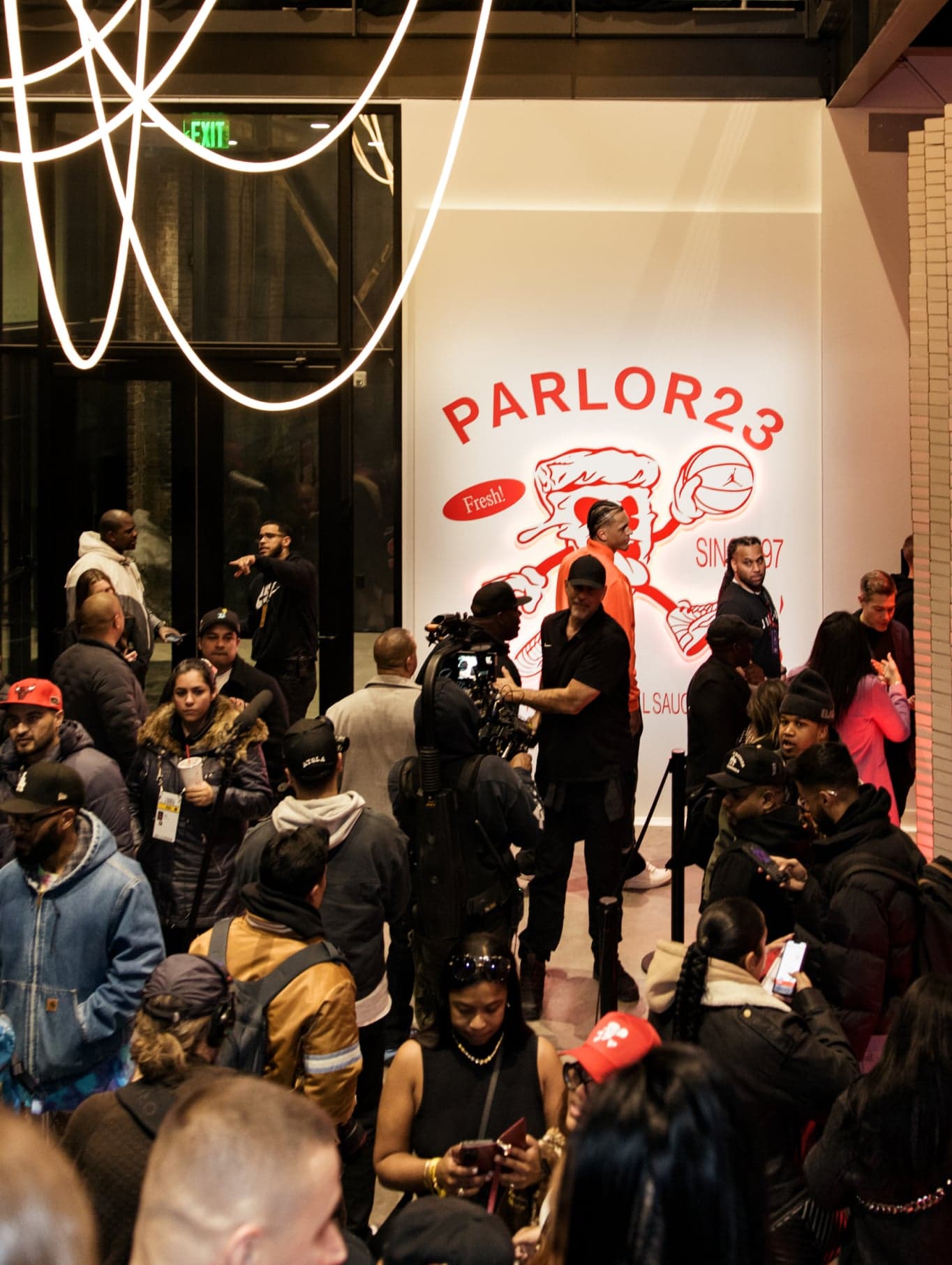 A crowd lining up past neon tubes toward the glowing Parlor 23 pizza mascot wall