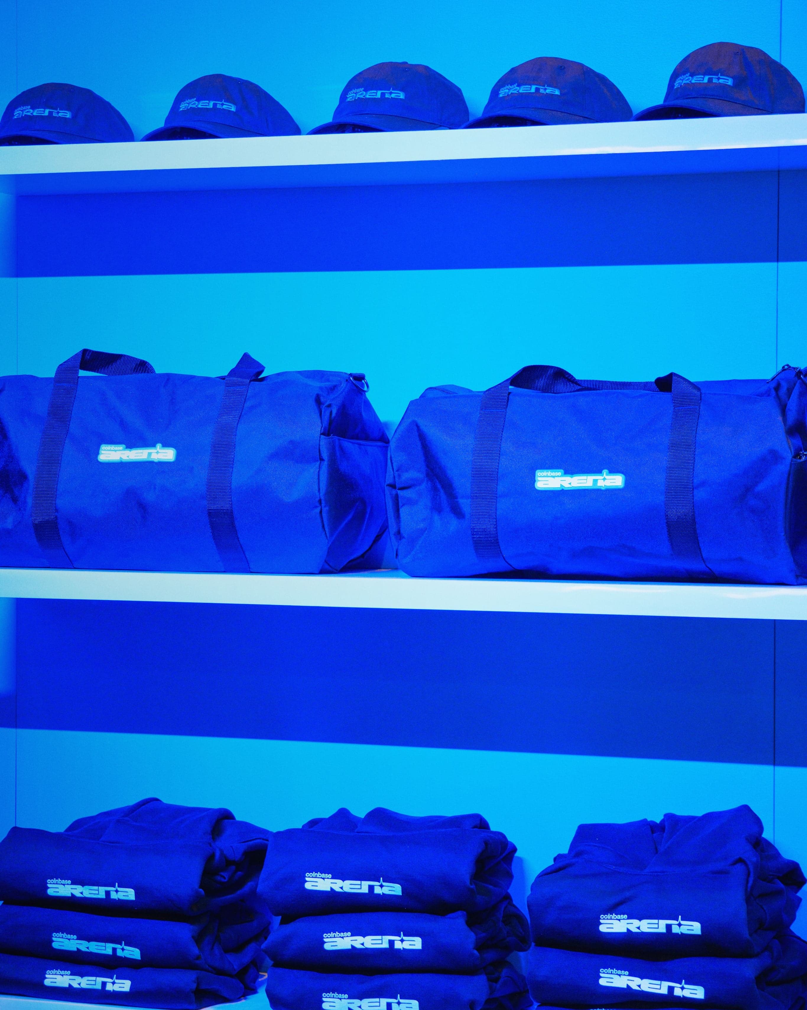 Shelves of Coinbase Arena caps, bags and folded merch under blue light