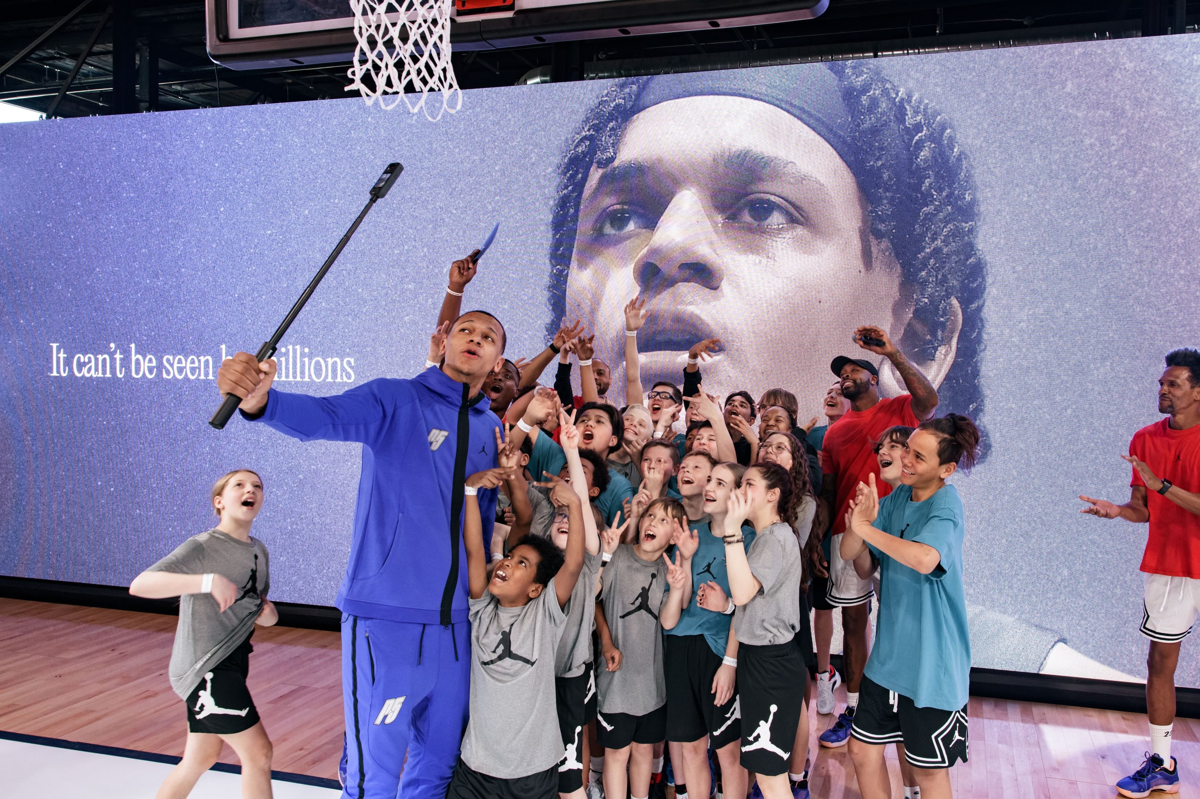A crowd of young players in Jordan shirts posing for a selfie under a hoop, in front of an LED wall with a player’s face