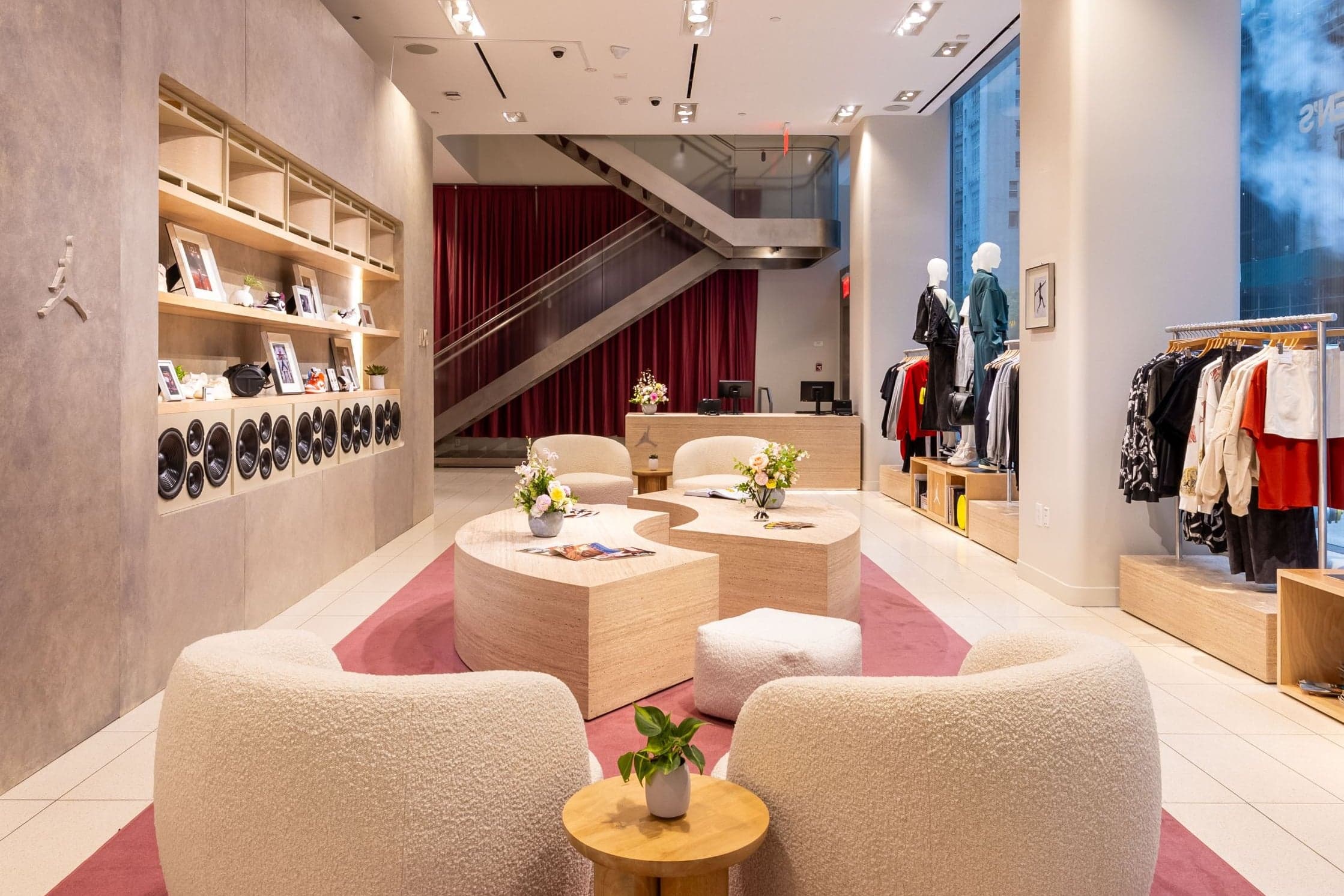 The lounge, with boucle armchairs and curved wooden tables on a pink rug, beside shelves of speakers and clothing rails