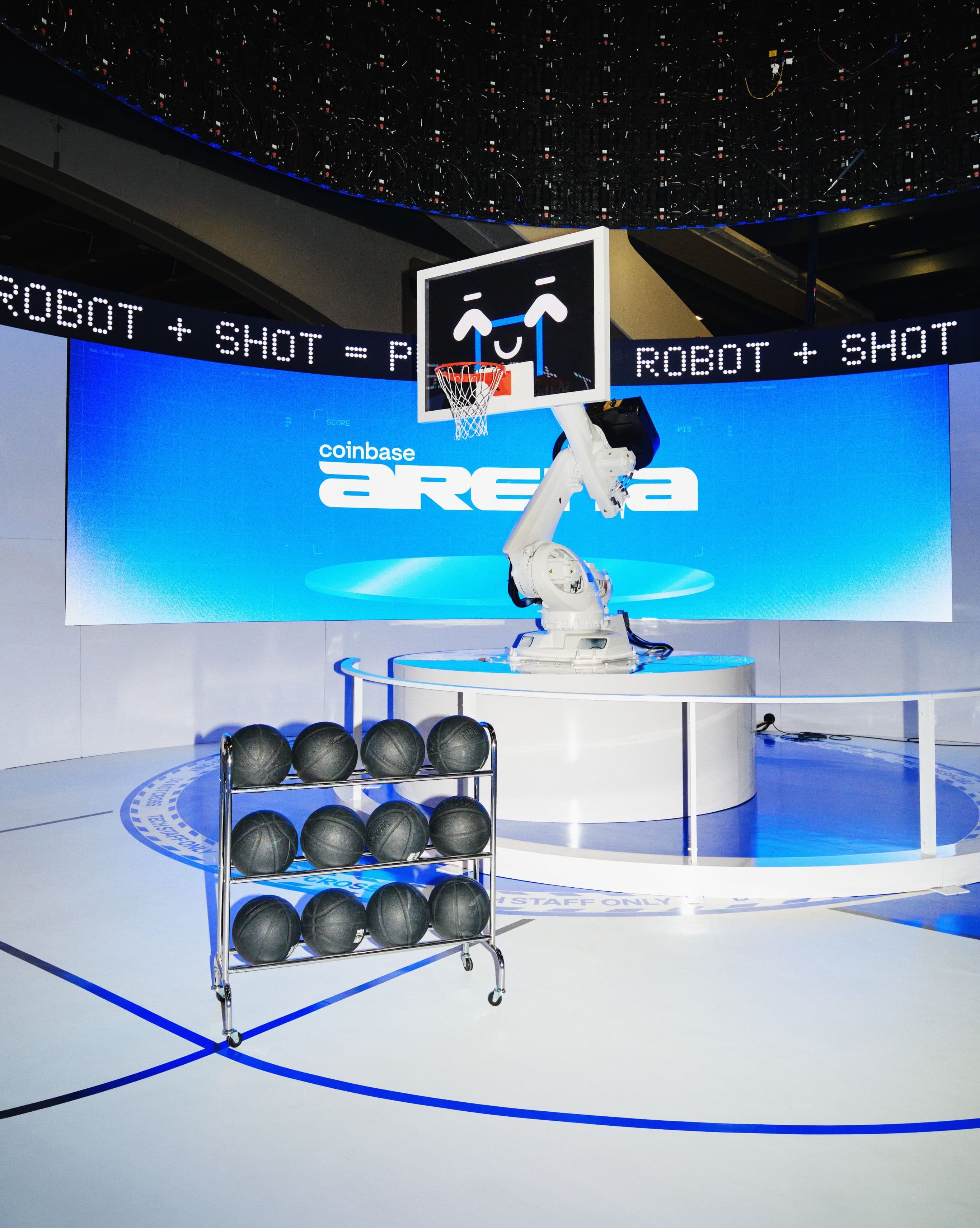 The robotic basketball hoop on its plinth beside a rack of basketballs, a “Robot + Shot” ticker running overhead