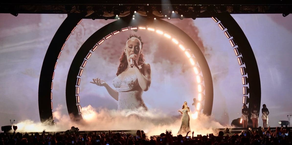 The singer on stage in front of a giant ring of lights, her face on the screen behind her amid smoke