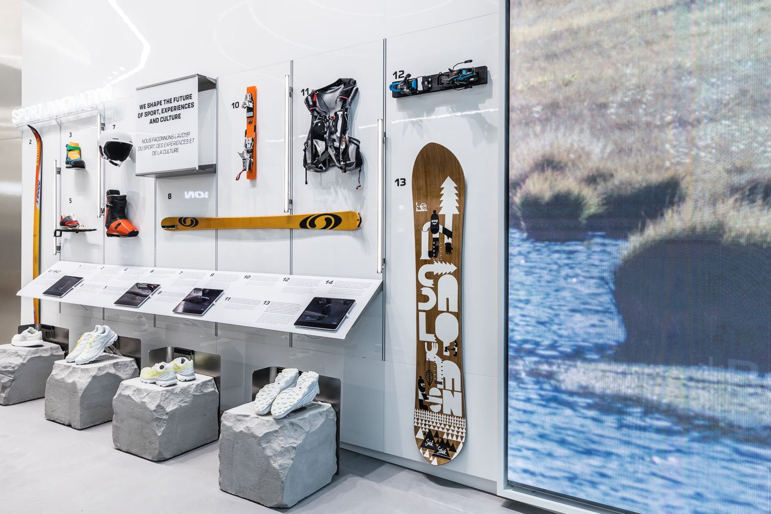 A snowboard and gear wall with shoes on stone plinths, beside a screen showing a river