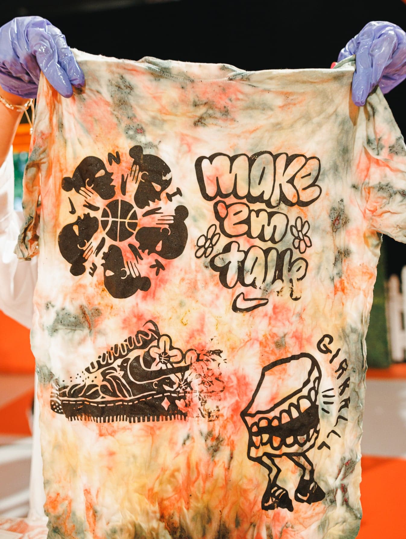 Hands holding up a tie-dye t-shirt printed with “Make ’em talk” and sketches of a sneaker and a grinning mouth