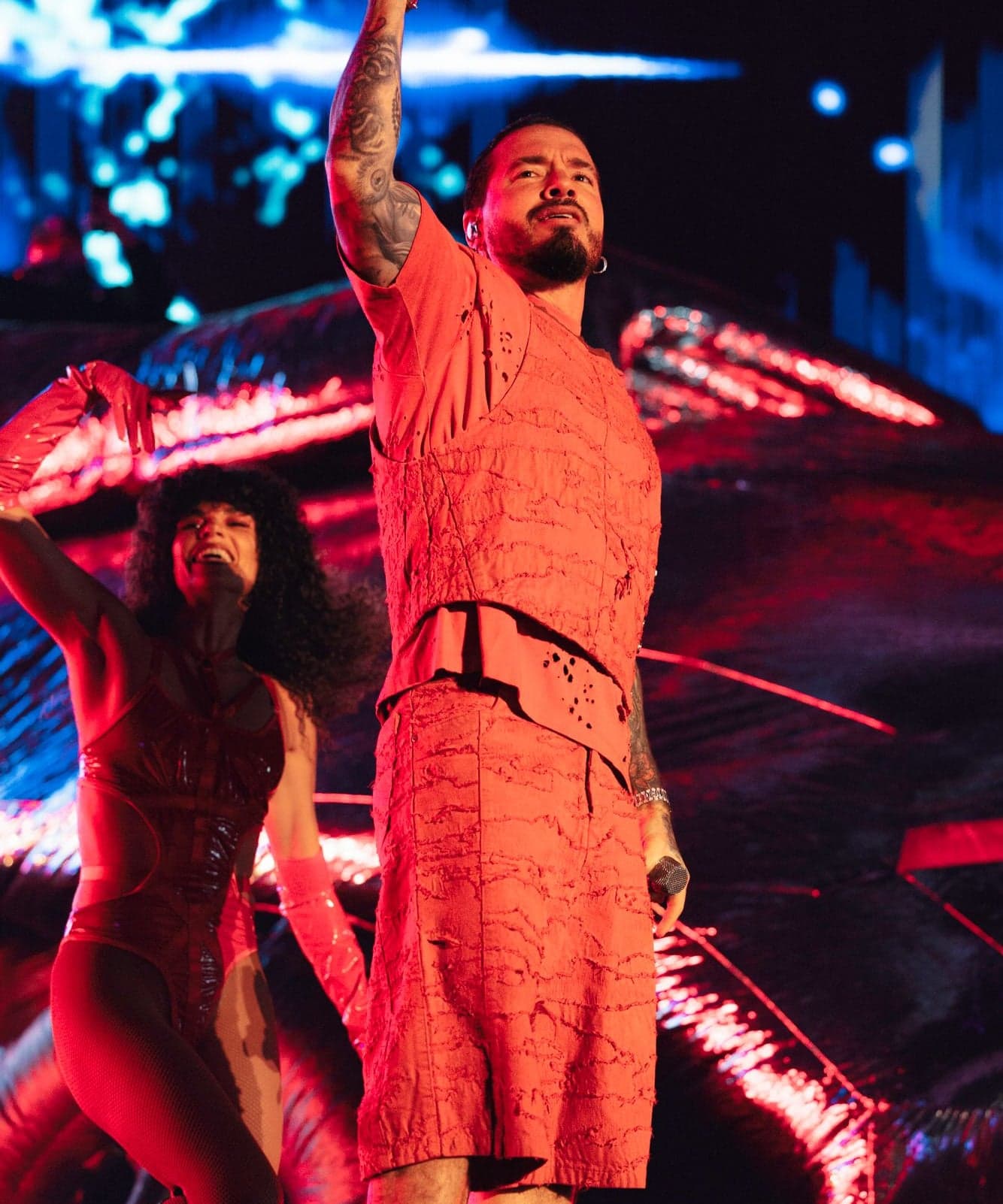 A performer in a terracotta outfit raising an arm on stage, with a dancer behind