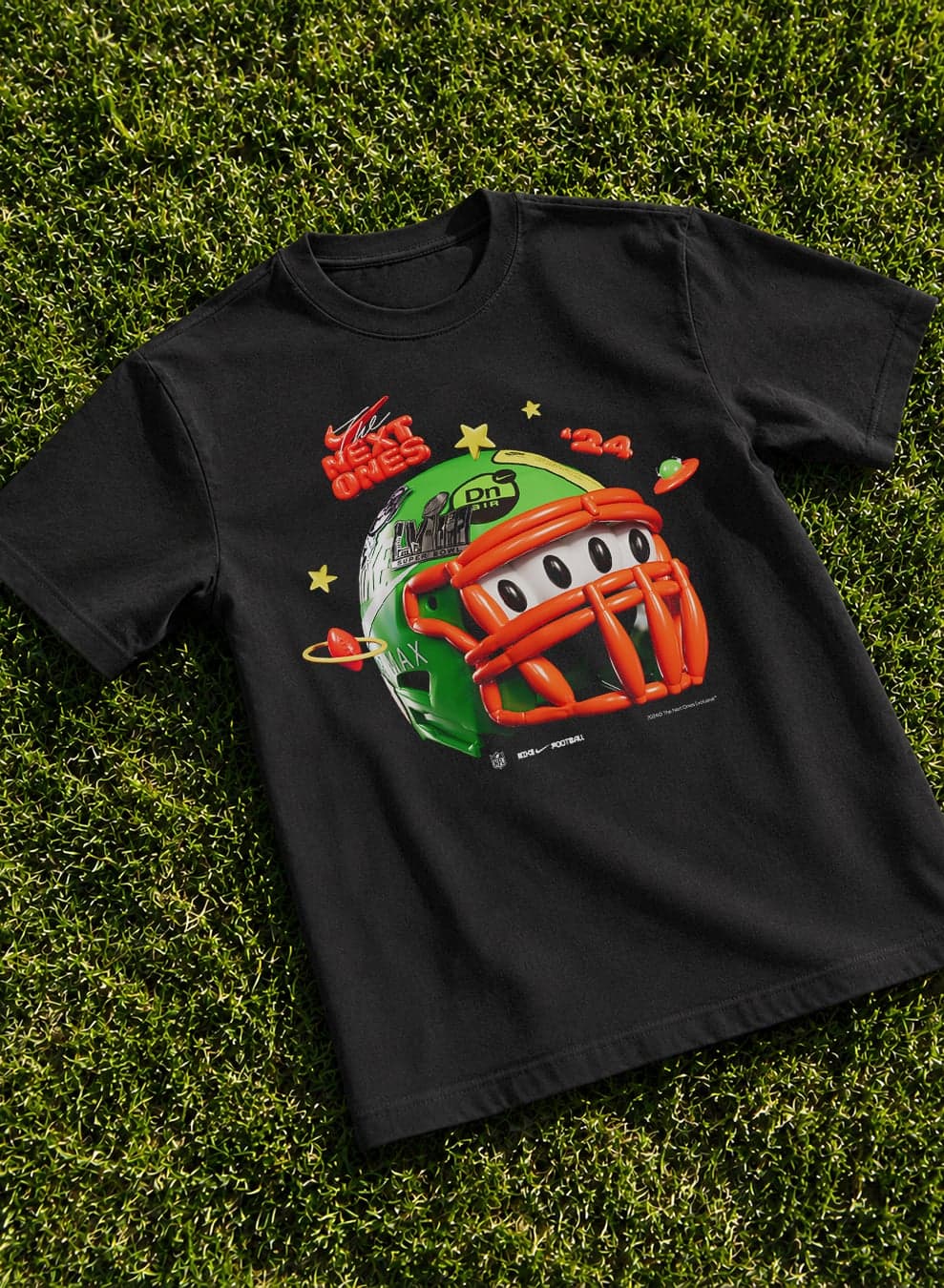 A black T-shirt printed with a cartoon green helmet and “The Next Ones” lying on grass