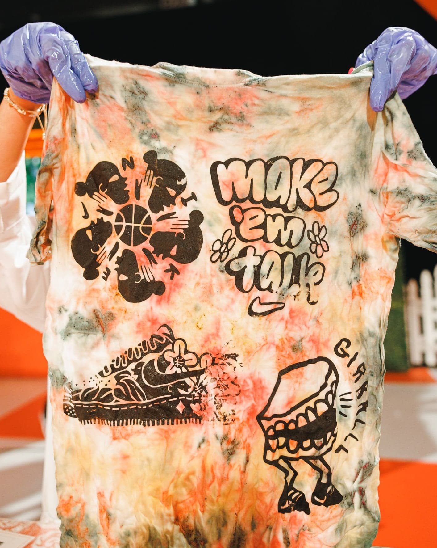 A tie-dye tee printed with Make ’Em Talk graphics, held up by gloved hands