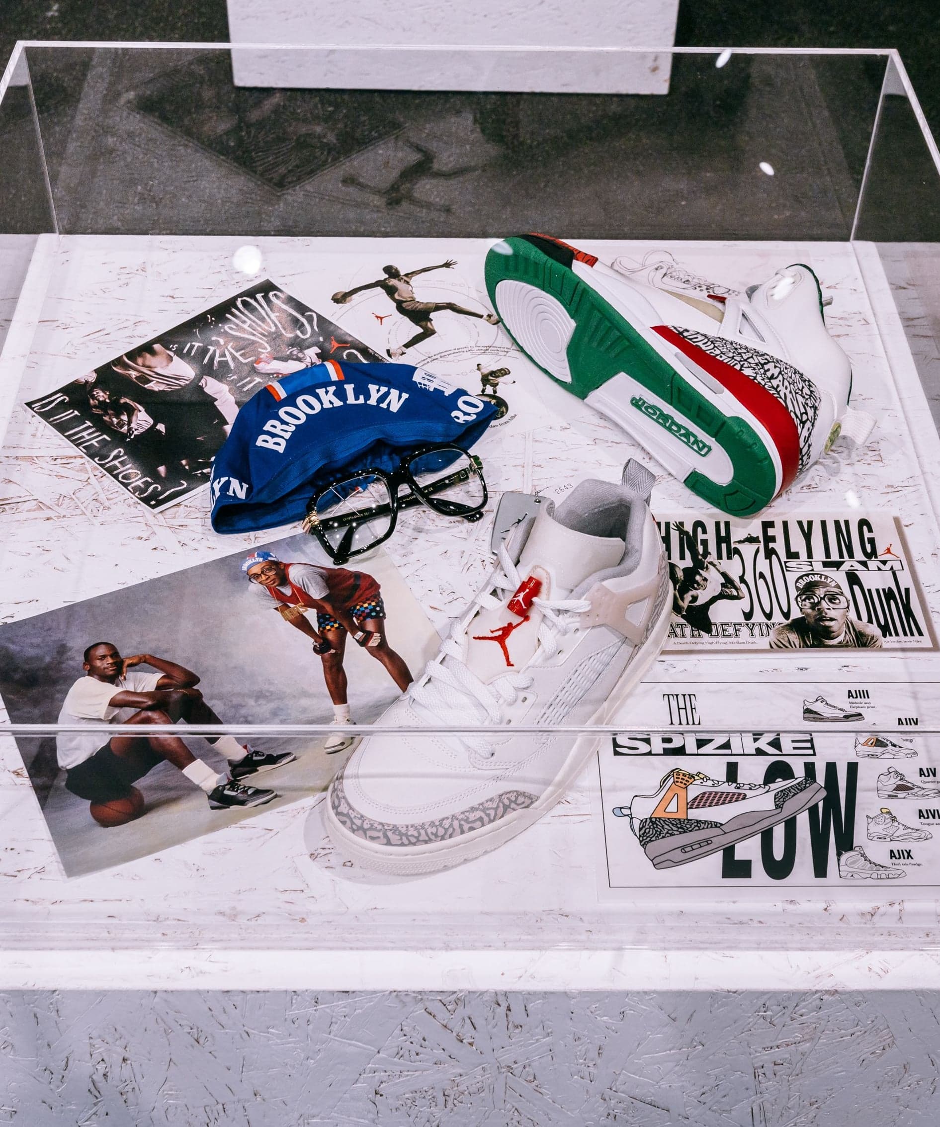 A glass display case holding Brooklyn sneakers, magazines and photos on a white plinth